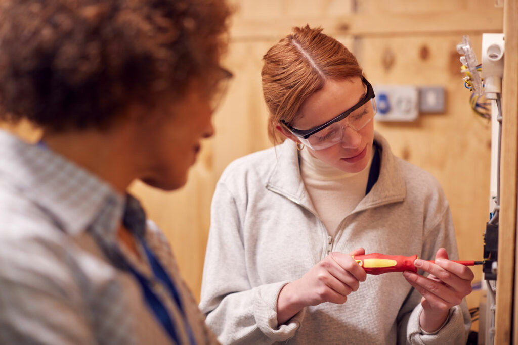 red head woman with safety goggles screws in a screw while a man watches on