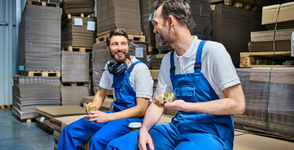 two young men sitting in a factory in their blue overalls enjoying sandwiches on a break