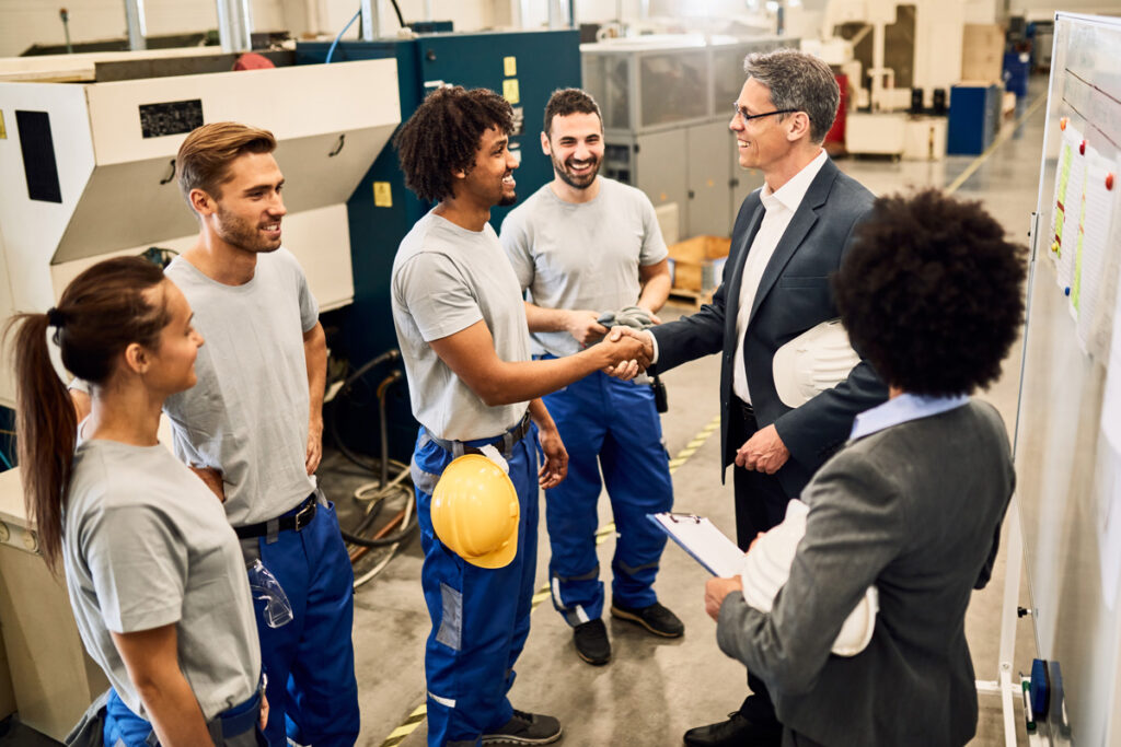 boss in a suit in a factory shakes hands with a young man while his team watches