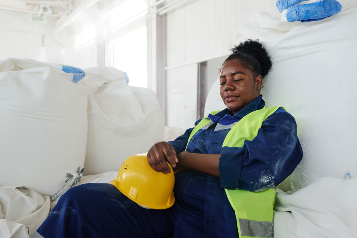 Sick female engineer in uniform sitting against huge white sack in workshop
