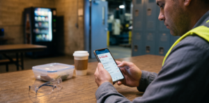 A photojournalistic, over-the-shoulder shot of a male production associate sitting at a worn wooden table in an industrial break room. He holds a smartphone showing a generic mobile banking app with an overdraft balance of -$133.00 in red text. In the soft-focus foreground lie folded safety glasses, a lunch container, and a coffee cup, with metal lockers and a plant floor visible in the background.