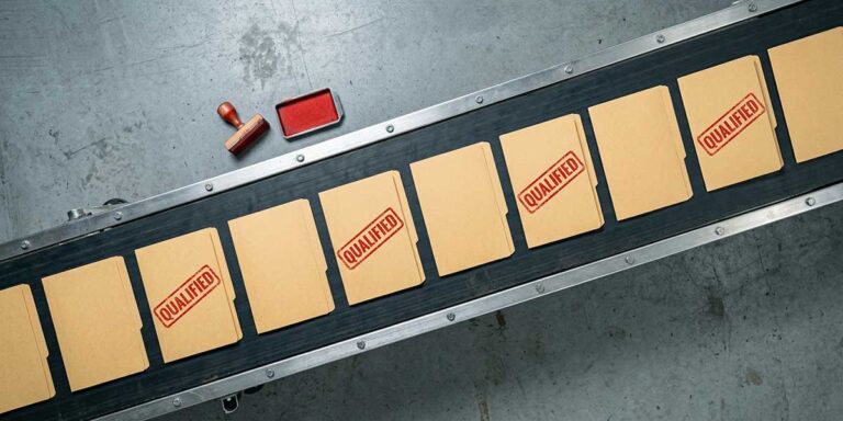 Top-down view of an industrial conveyor belt carrying ten manila job folders, four stamped “QUALIFIED” in red, with a red ink pad and wooden stamp beside the belt.
