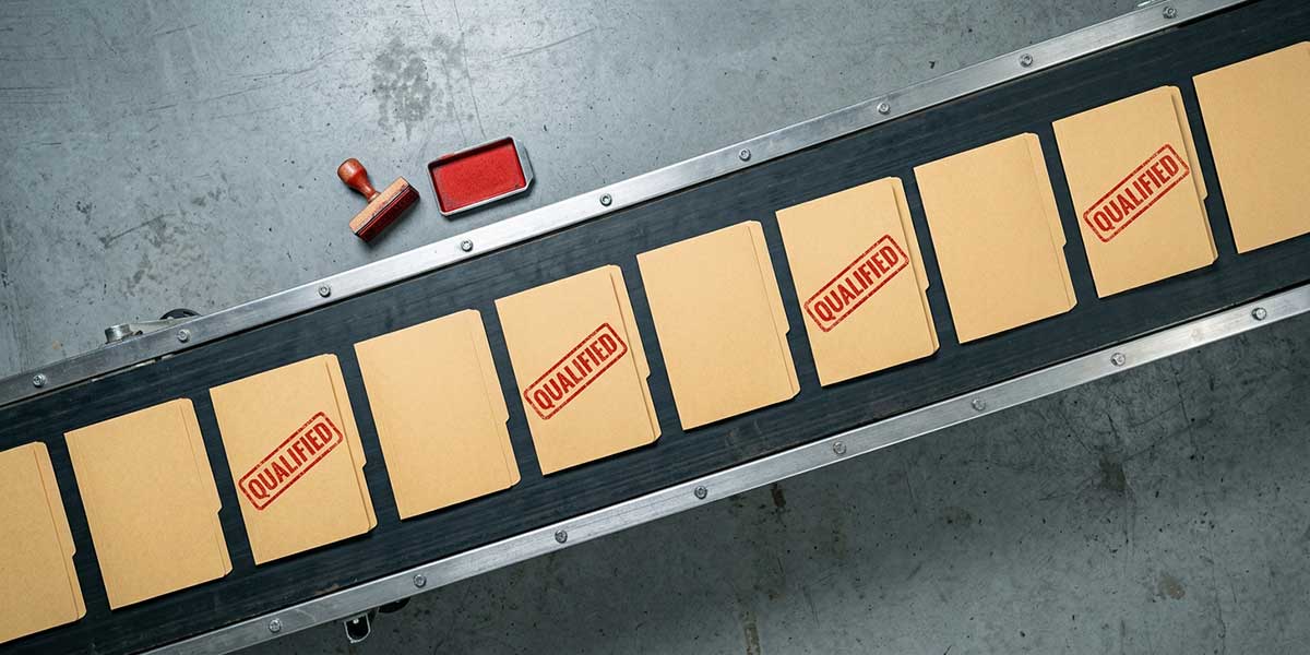 Top-down view of an industrial conveyor belt carrying ten manila job folders, four stamped “QUALIFIED” in red, with a red ink pad and wooden stamp beside the belt.